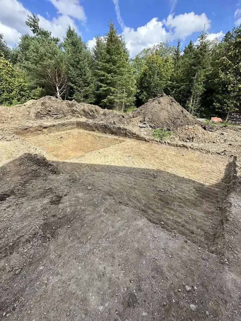 Excavated building pad cut into a treed lot with graded soil and spoil piles