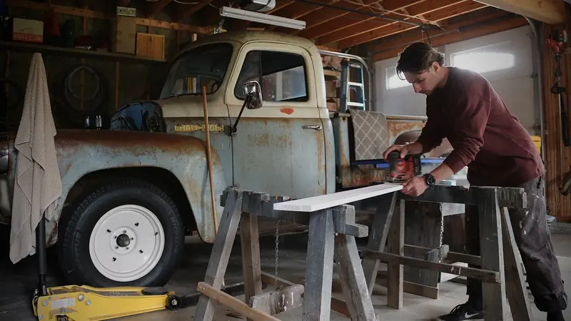 Blake Cole sanding a board on sawhorses in his workshop beside a vintage pickup truck
